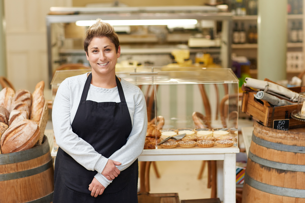 person in apron poses next to bread and tarts in a showcase