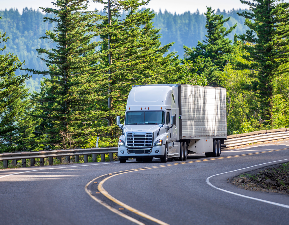 tractor trailer drives up winding road with pine trees in the background