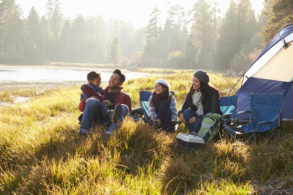 family sits in camping chairs next to a tent by a body of water